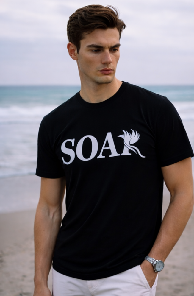 Male model wearing a black SOAR logo t-shirt standing on a beach with ocean waves in the background.