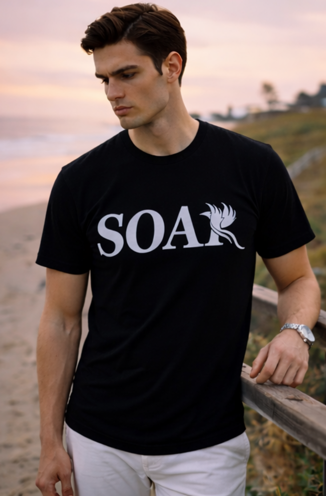 Male model wearing a black SOAR logo t-shirt leaning on a wooden railing at the beach during sunset.
