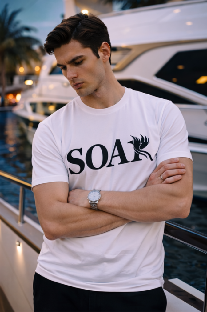 White SOAR logo t-shirt on male model with arms crossed, photographed at a marina with luxury yachts in the background.